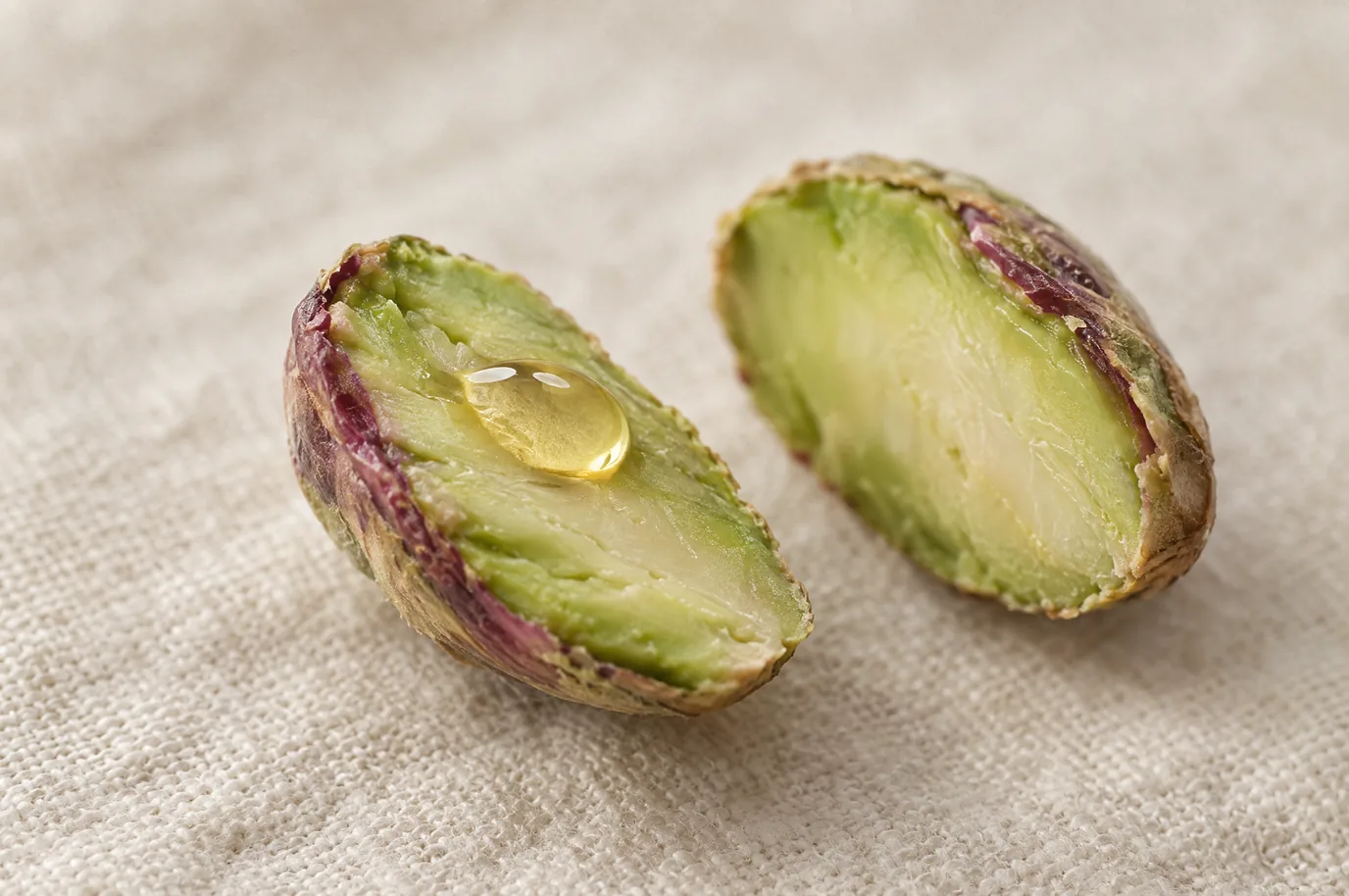 A single pistachio kernel split in half on cream linen, a bead of pale gold oil resting on the cut face.
