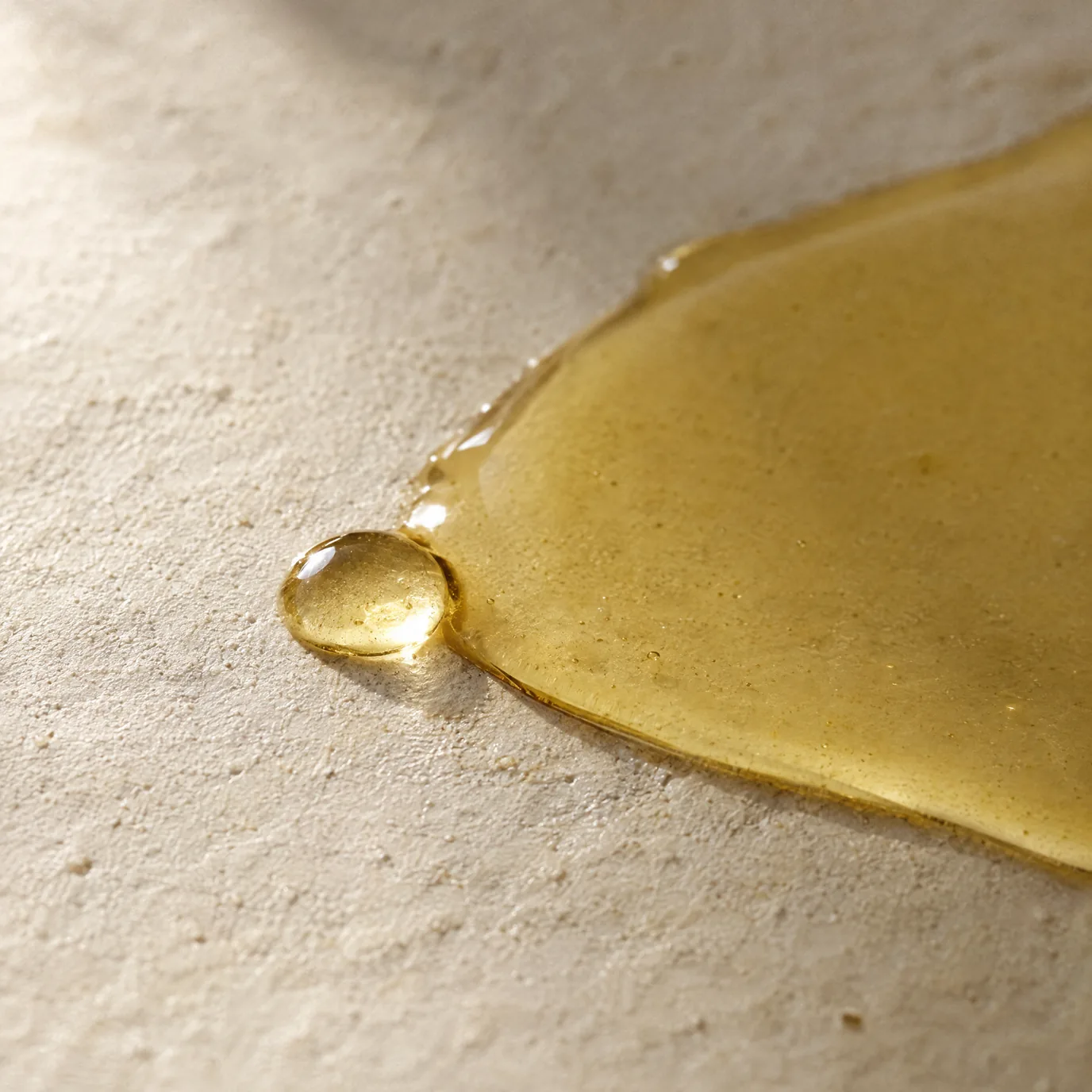 A pool of pale golden oil on cream plaster, a single drop at its edge.